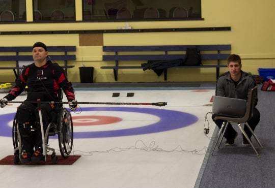 Brock crouches at computer beside paralympic curling athlete sitting biomechanical wheelchair on the ice