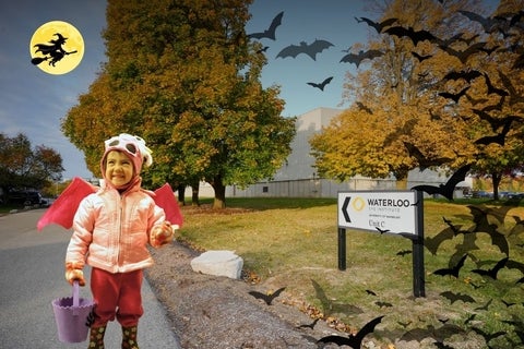 Child in costume next to Waterloo Eye Institute sign. Bats and a witch are in the background.