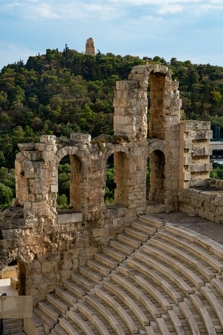 View of the Odeon of Herodes Atticus on the Acropolis in Athens, showing the stone seating and facade of the ancient Roman theater with a wooded hill in the background.