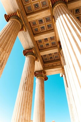 Upward view of ancient Greek Doric columns in Athens, showing ornate painted details on the coffered ceiling against a bright blue sky.