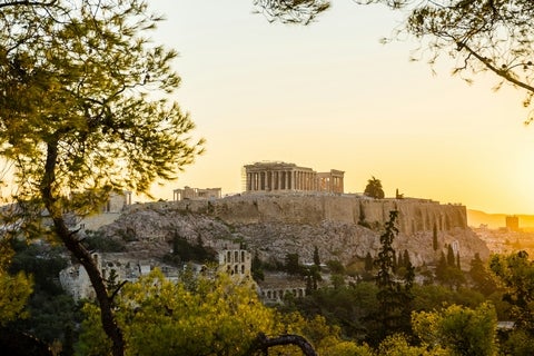 View of the Acropolis of Athens and the Parthenon at sunset, framed by trees in the foreground.