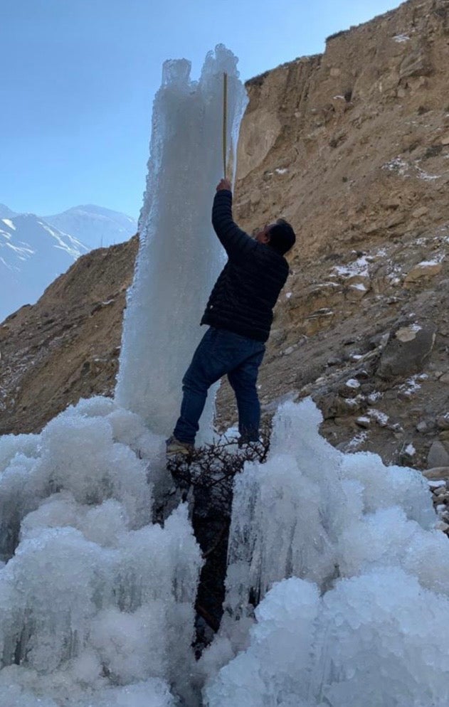 A man stands on a base of frozen ice and reaches up to adjust a vertical pipe extending from a tall, cylindrical ice tower (an ice stupa). The scene is set in a dry, steep mountain valley with a rocky slope in the background 