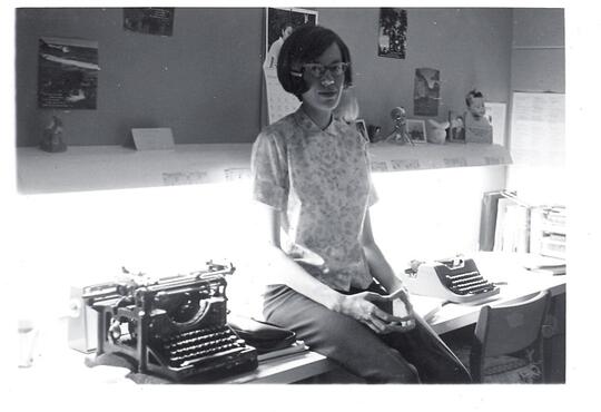 black and white photo of a young female student sitting on a desk beside an old typewritter