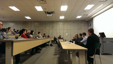 A WatITis presentation in progress.  A row of presenters are sitting in a line at the front of a classroom, and participants are listening to them from the classroom seats.