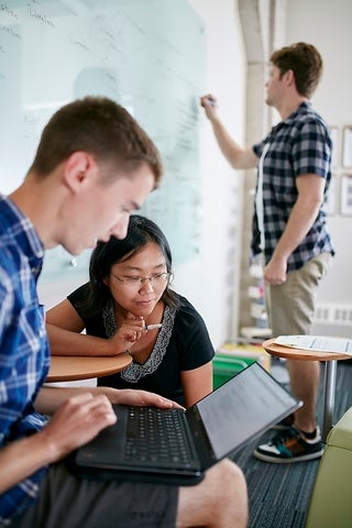 A professor with two students. One students is looking at a laptop while the other is writing on a chalkboard.