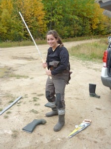 A woman holding a long stick wearing hip waders.