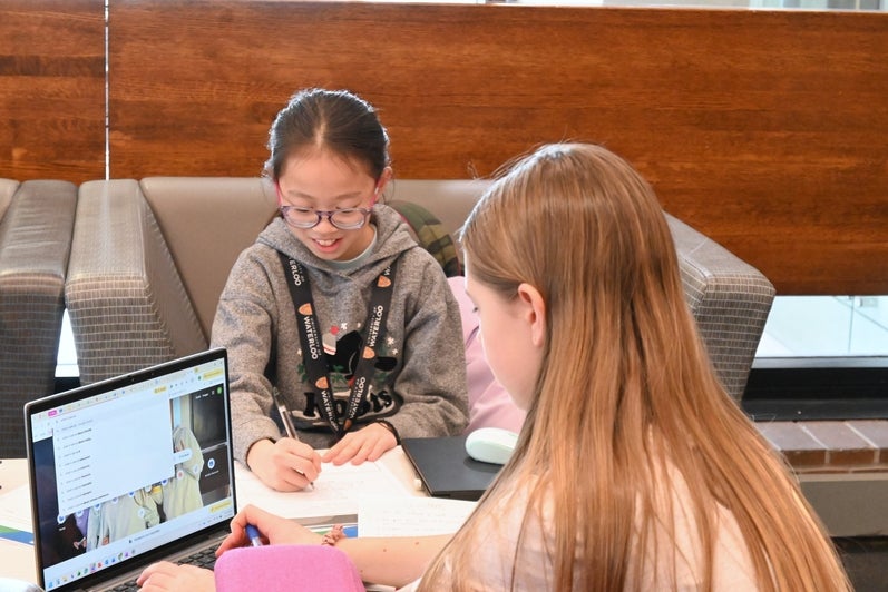 2 girls working at a table, one is smiling which writing on a piece of paper, the other you see the back of her head while she is googling something on a laptop