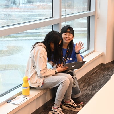 two girls sharing one computer sitting in front of a window. One of the girls is looking at the camera and waving