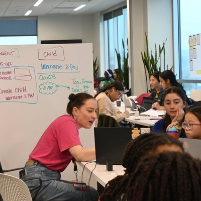 2 Volunteers helping a participant with their project. A whiteboard with a wireframe of the app is in the background