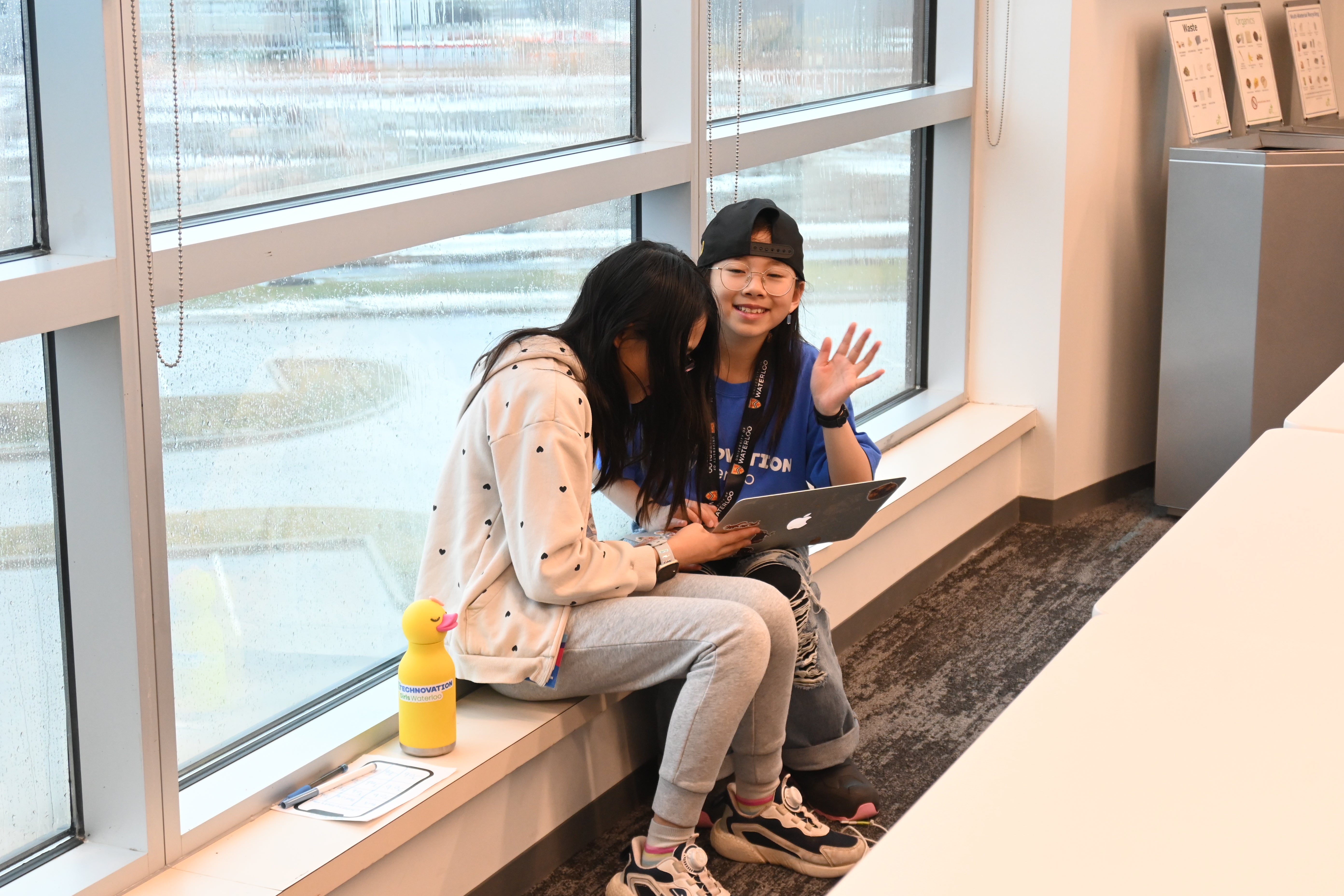 two girls sharing one computer sitting in front of a window. One of the girls is looking at the camera and waving