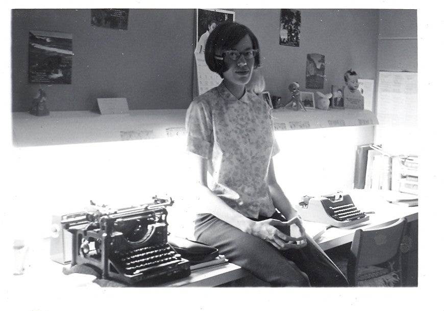 black and white photo with a woman sitting on a desk beside a typewriter