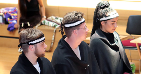 Three students standing side by side wearing black cloaks and black and white headbands