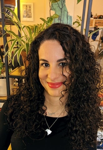 Photo of Amanda Fogaça. She has long dark curly hair and dark brown eyes. She is sitting in front of houseplants, wearing a black sweater, silver necklace, and red lipstick. She is looking into the camera and smiling.