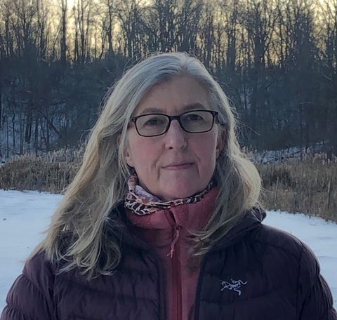 Headsot of Liz Newbery. She has shoulder length light coloured hair and is wearing black rimmed glasses. She is standing outside in front of a forest in the winter.