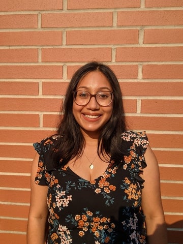 Sarah Menzies. She has long dark hair and is wearing glasses. She is standing in front of a red brick wall, looking into the camera, and smiling.