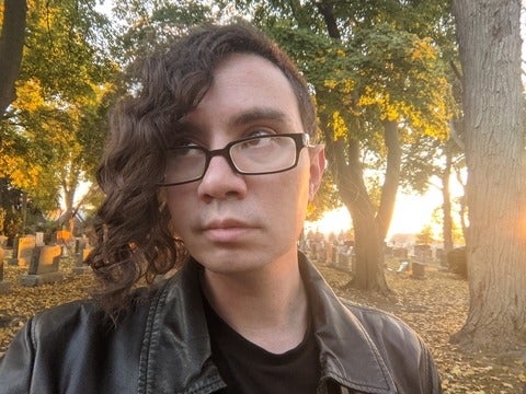 Chris Lawrence has short brown hair with long wavy bangs and brown eyes. They are wearing black rimmed glases and a black leather jacket and standing with their back to a graveyard during a sunset. They are looking away from the camera.