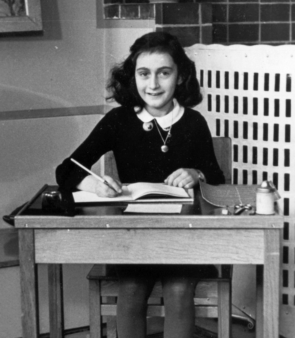 An image of Anne Frank writing at a desk and smiling