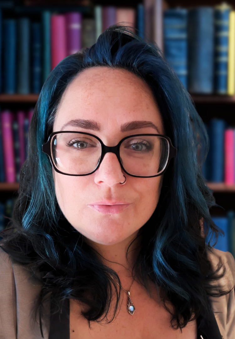 Photo of Meghan Ashdown. She has long black and blue hair and is wearing black rimmed glasses and a black top. She is looking into the camera and there is a library with colourful books in the background.