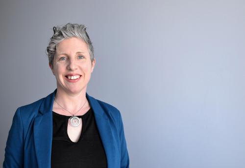 Image of Clare Bermingham. She has short grey hair and is wearing a black blouse and blue blazer. She is standing against a grey background and smiling directly at the camera.