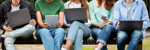 Group of young people spending time with electronic devices outdoors, sitting on bench in park with gadgets in hands