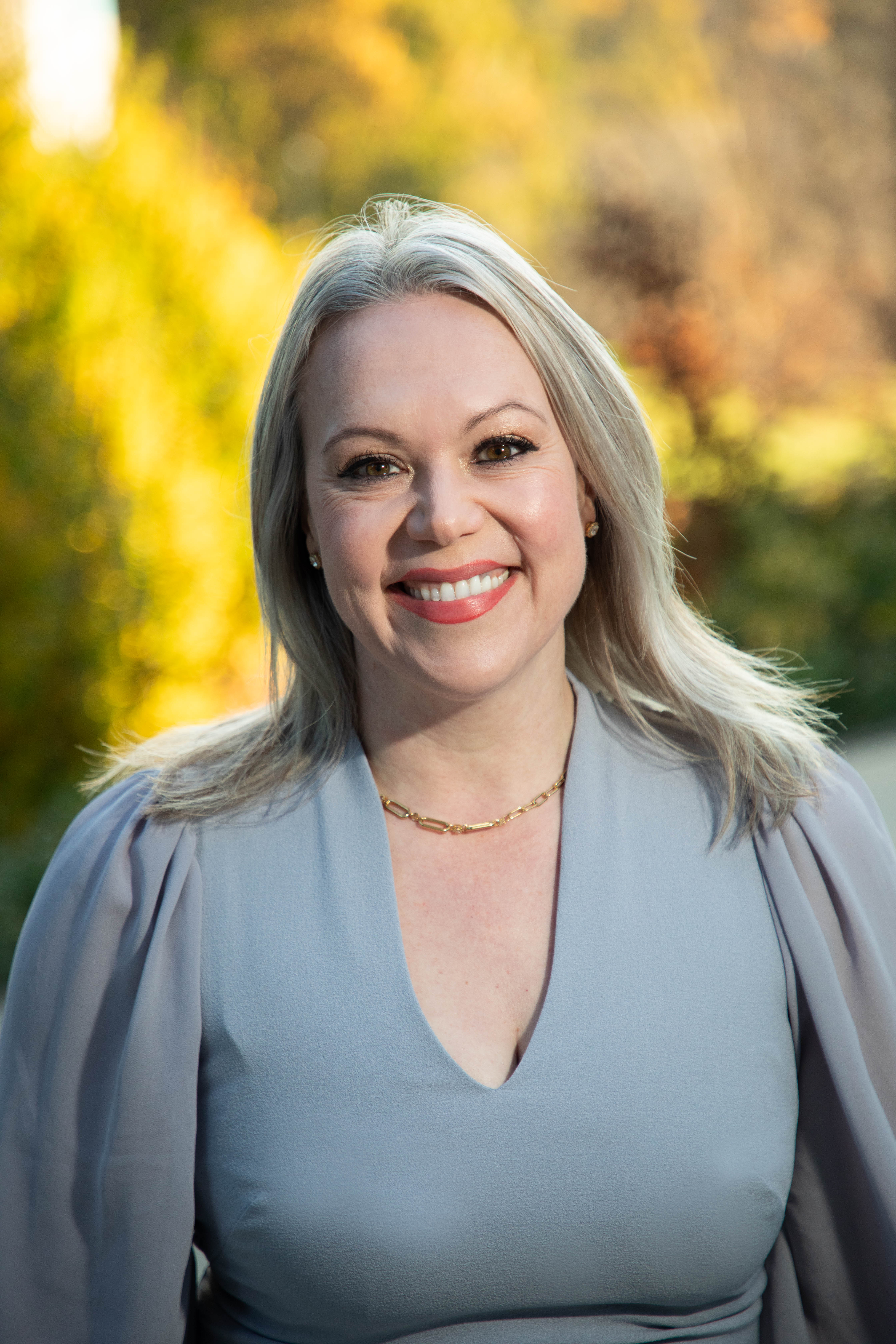 Photo of Anne Fannon, wearing a grey dress, set against a blurred autumn outdoor background.