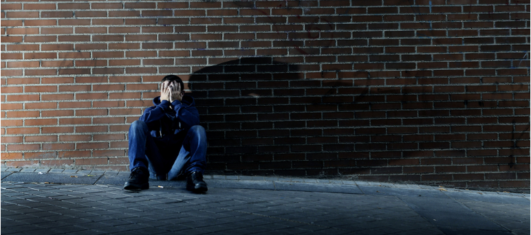 Young person sitting against a brick wall with their hands over their face